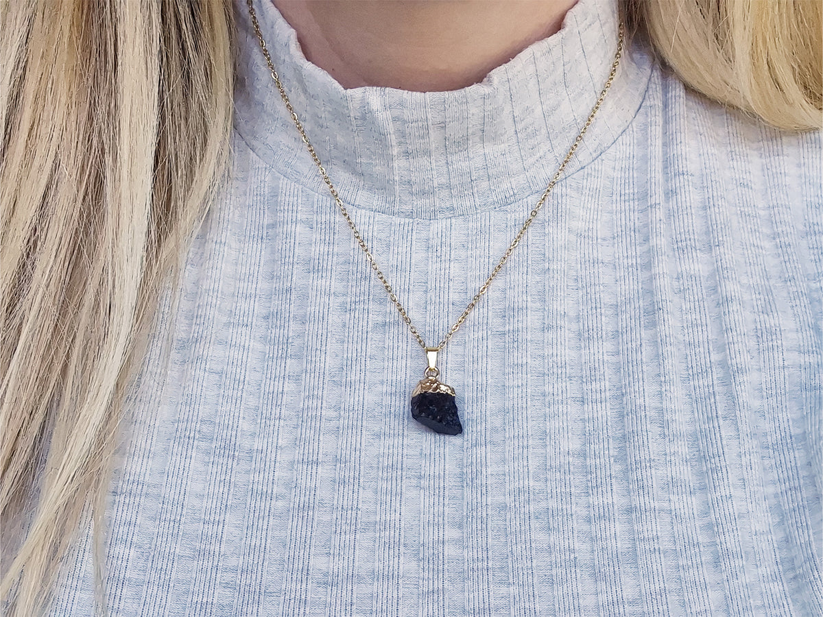 Woman wearing a gold-plated black tourmaline necklace with a raw crystal pendant over a light blue grey top.